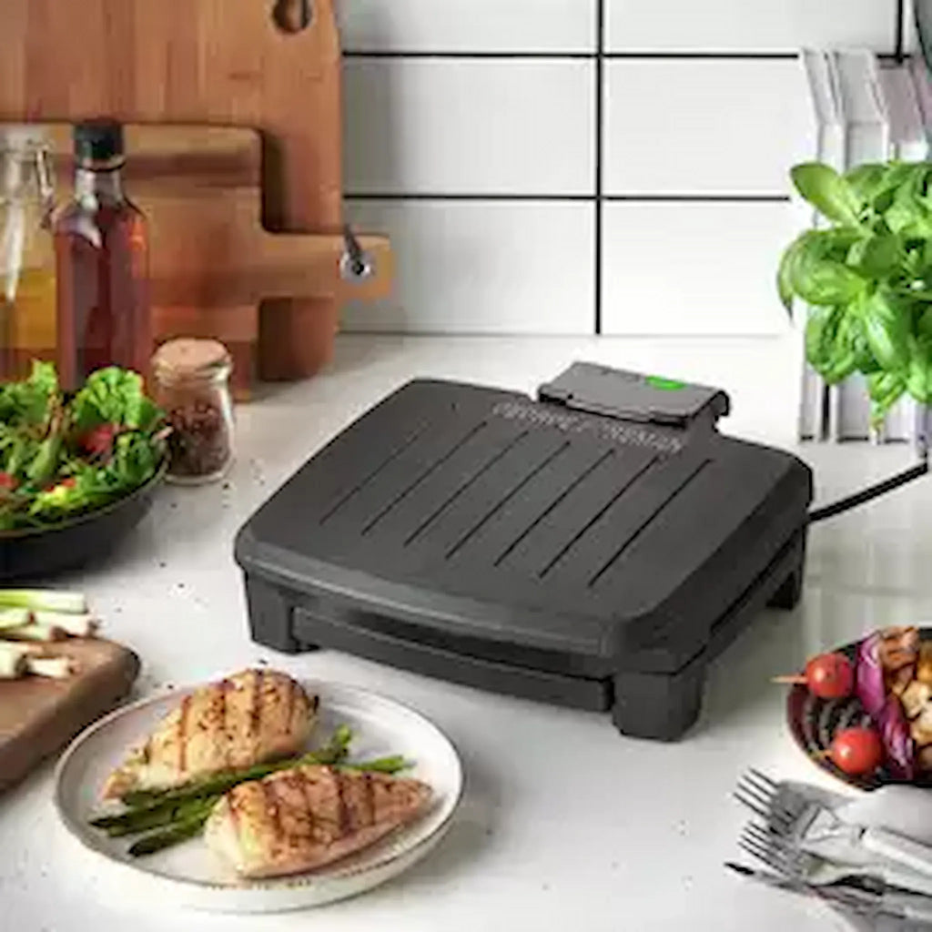A black George Foreman Immersa grill on a kitchen countertop with food being cooked on it, surrounded by various kitchen items and a plate with grilled food in front.